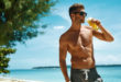 Uomo abbronzato in costume con gli occhiali da sole beve una bevanda alla frutta su una spiaggia