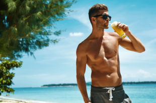 Uomo abbronzato in costume con gli occhiali da sole beve una bevanda alla frutta su una spiaggia