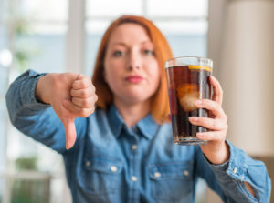 Giovane donna tiene in mano bicchiere con bevanda zuccherata tipo cola con ghiaccio e limone mentre con l'altra mano fa il segno del pollice in giù