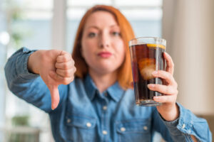 Giovane donna tiene in mano bicchiere con bevanda zuccherata tipo cola con ghiaccio e limone mentre con l'altra mano fa il segno del pollice in giù