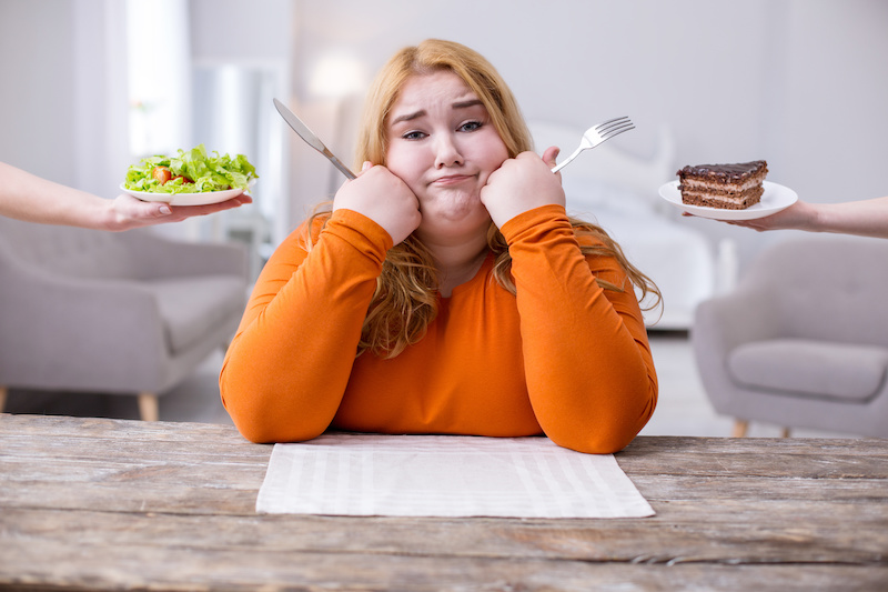 Ragazza in sovrappeso seduta a tavola con forchetta e coltello mentre due mani femminili le porgono un'insalata e una fetta di torta; concept: dieta, obesità