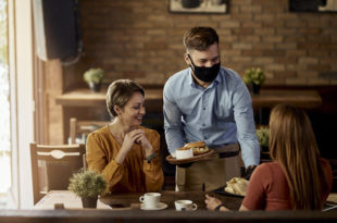Happy waiter wearing protective face mask while serving food to guests in a restaurant.