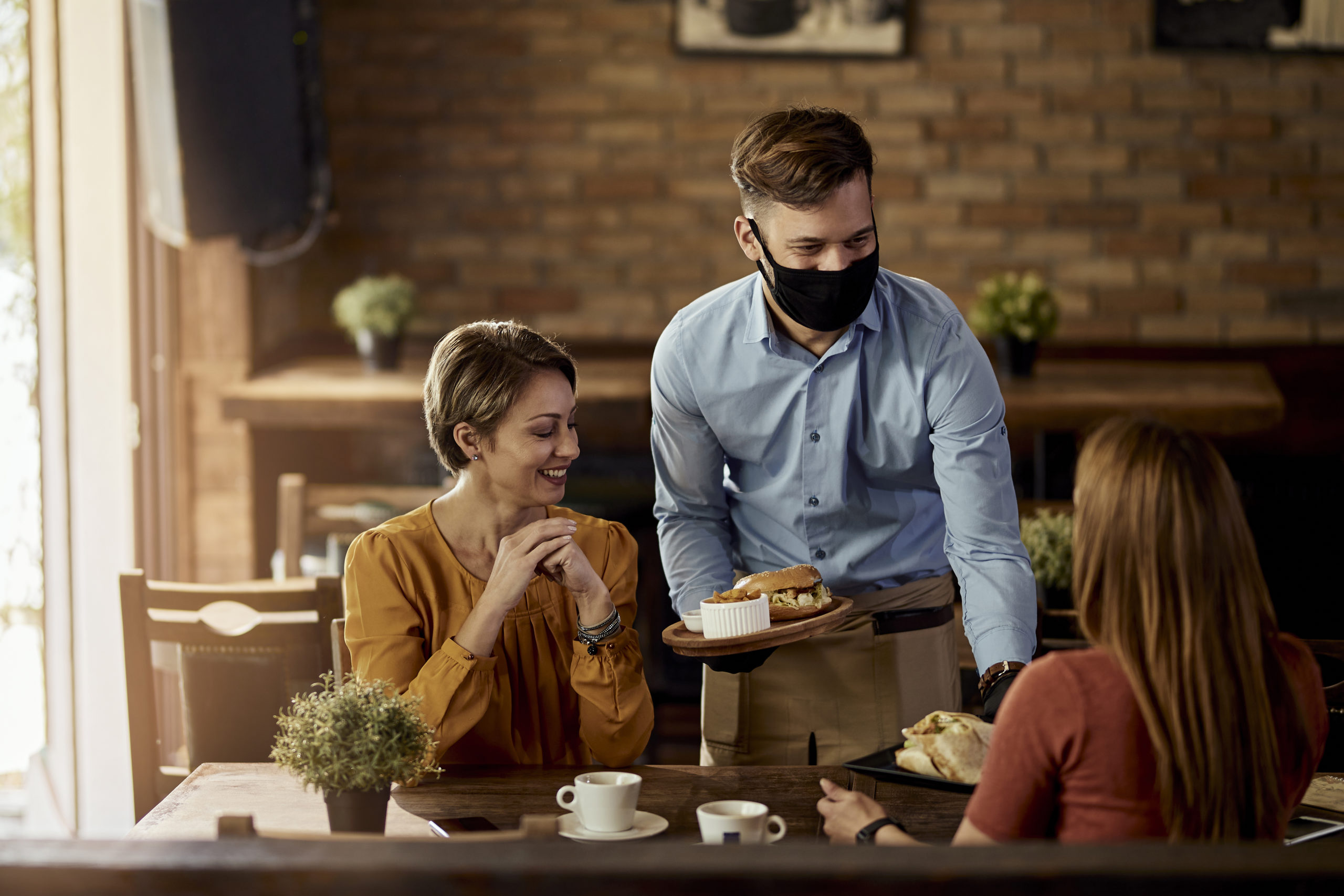Happy waiter wearing protective face mask while serving food to guests in a restaurant.
