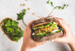 Panino con burger vegetale, insalata e verdure tenuto da due mani, sullo sfondo verdure in foglia e ceci o soia