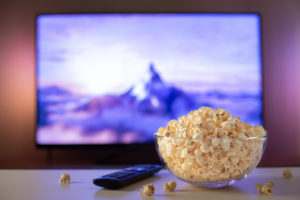 Ciotola di vetro piena di popcorn accanto a telecomando; sullo sfondo televisione con film o programma tv