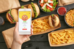 Smartphone con aperta app di food delivery; sullo sfondo contenitori per asporto o consegna a domicilio con hamburger, patatine, salse e insalata
