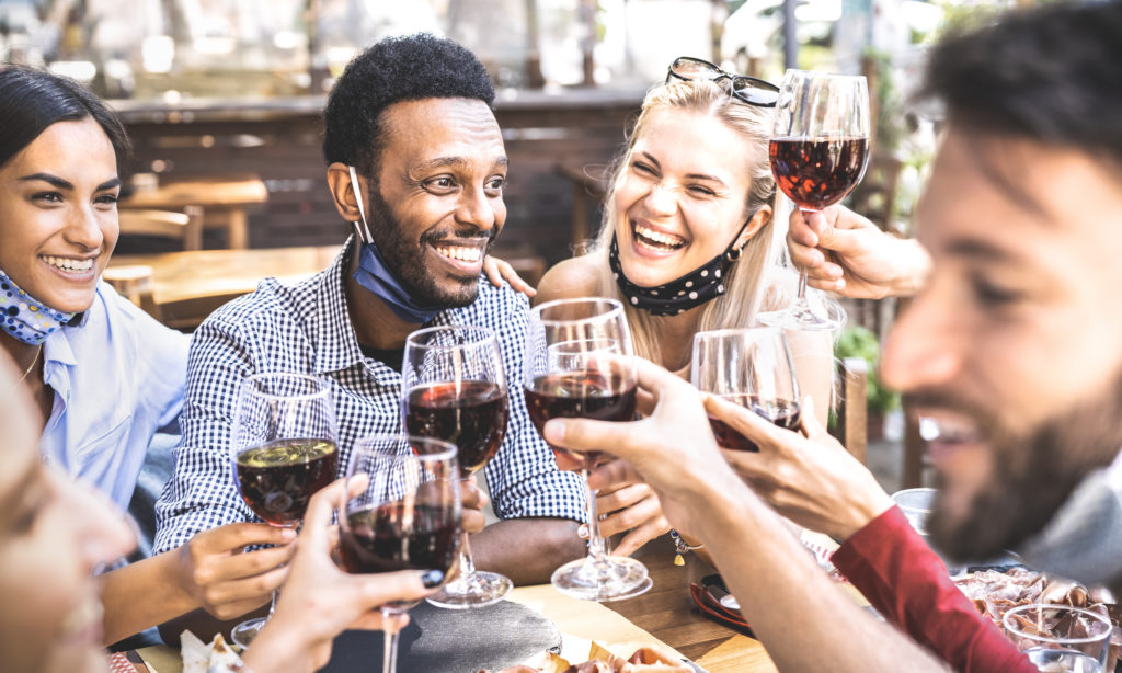 Friends toasting red wine at outdoor restaurant bar with open face mask - New normal lifestyle concept with happy people having fun together on warm filter - Focus on afroamerican guy