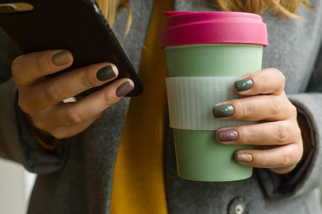 Una donna con smalto multicolore tiene in mano una tazza riutilizzabile in bambù con caffè o altra bevanda calda; nell'altra mano tiene uno smartphone