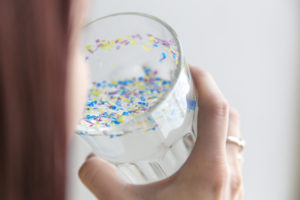 Donna di spalle beve bicchiere pieno di frammenti di plastica; concept: microplastiche