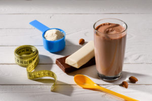 Prodotti protein/proteici: bicchiere con bevanda al cioccolato o caffè o nocciola, barrette, polvere in misurino blu, cucchiaino arancione e metro