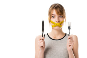 Donna con forchetta e coltello in mano e la bocca chiusa da un metro a nastro; concept: dieta