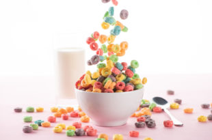 Cereali da colazione multicolore ricadono in una tazza, altri sono sparsi intorno sul tavolo; sulla destra un cucchiaio e sullo sfondo un bicchiere di latte