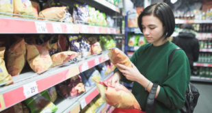 Giovane donna con cestino sotto braccio sceglie chips dagli scaffali degli snack del supermercato
