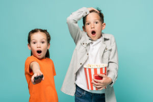 Un bambino con secchiello di popcorn e una bambina con telecomando ed espressione stupita; concept: tv, cinema, televisione
