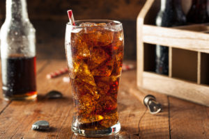 Bicchiere di bevanda zuccherata tipo cola con ghiaccio e cannuccia; sul retro bottiglie di vetro con la stessa bevanda