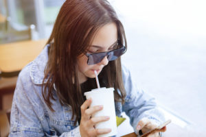 Ragazza adolescente beve una bevanda zuccherata con una cannuccia mentre guarda lo smartphone adolescenti