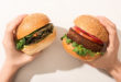 A sinistra una mano tiene un panino con un burger vegetale e insalata, a destra una mano tiene un panino con un diverso tipo di burger vegetale, insalata e pomodoro