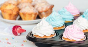 Cupcake nello stampo con glassa bianca, rosa e azzurra; sullo sfondo un piatto con cupcake senza glassa; accanto un sac à poche e perline di zucchero sparse