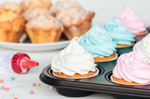Cupcake nello stampo con glassa bianca, rosa e azzurra; sullo sfondo un piatto con cupcake senza glassa; accanto un sac à poche e perline di zucchero sparse