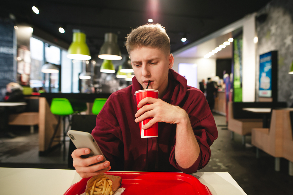 ragazzo beve coca-cola mentre guarda smartphone in ristorante fast food maxi porzioni