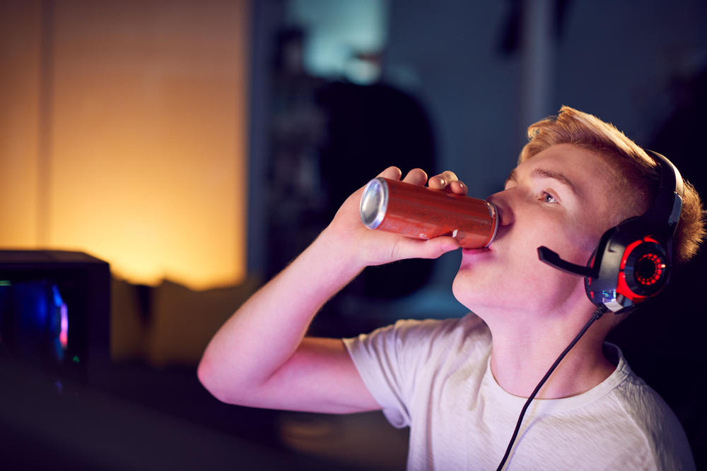 Ragazzo adolescente con cuffie da gamer beve un energy drink o una bevanda zuccherata da una lattina