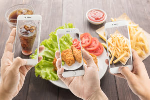 Tre smartphone inquadrano, da sinistra a destra, bicchiere di bibita gassata, pollo fritto e patatine fritte; concept junk food e influencer