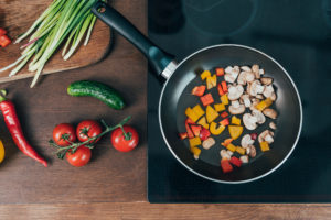 Padella con verdure e funghi a tocchetti su piastra a induzione, accanto a pomodori, peperoncini, cetriolo e erba cipollina