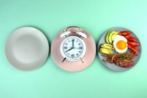 Tre piatti: a sinistra vuoto, al centro con una sveglia, a destra pieno di cibo; concept: digiuno intermittente