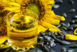 Bicchiere di olio di girasole in primo piano davanti a due fiori di girasole, con semi di girasole sparsi su superficie di ardesia