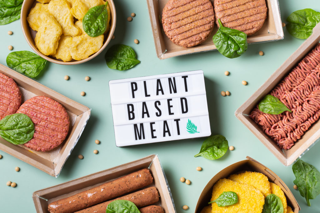 Cartello con la scritta “plant based meat” circondato da assortimento di prodotti vegetali – burger, salsicce, nuggets, cotolette – semi di soia e foglie di basilico