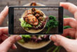 Smartphone tenuto da mani femminili inquadra piatto di carne di maiale con insalata e funghi, accanto al piatto mazzo di prezzemolo o coriandolo; concept social media