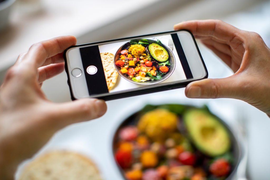 Smartphone tenuto da mani femminili inquadra bowl vegana con avocado e verdure