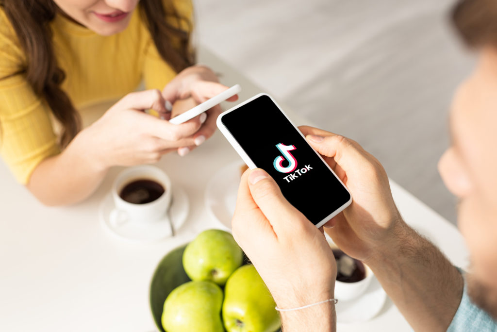 Un uomo e una donna guardano i propri smartphone seduti a un tavolino con tazze di caffè e ciotola di mele; sullo smartphone dell'uomo è presente il logo del social media TikTok