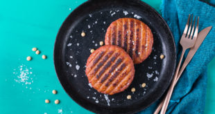 Due burger vegetali cotti alla piastra appoggiati su un piatto nero su superficie turchese; sulla destra tovagliolo blu, forchetta e coltello; sparsi semi di soia e granelli di sale