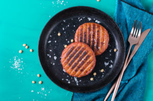 Due burger vegetali cotti alla piastra appoggiati su un piatto nero su superficie turchese; sulla destra tovagliolo blu, forchetta e coltello; sparsi semi di soia e granelli di sale