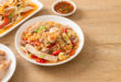 Piatto di noodles con verdure, gamberi, pollo e chili, circondato sulla sinistra da piatto con petto di pollo a fette, piatto con verdura mista e ciotolina di salsa