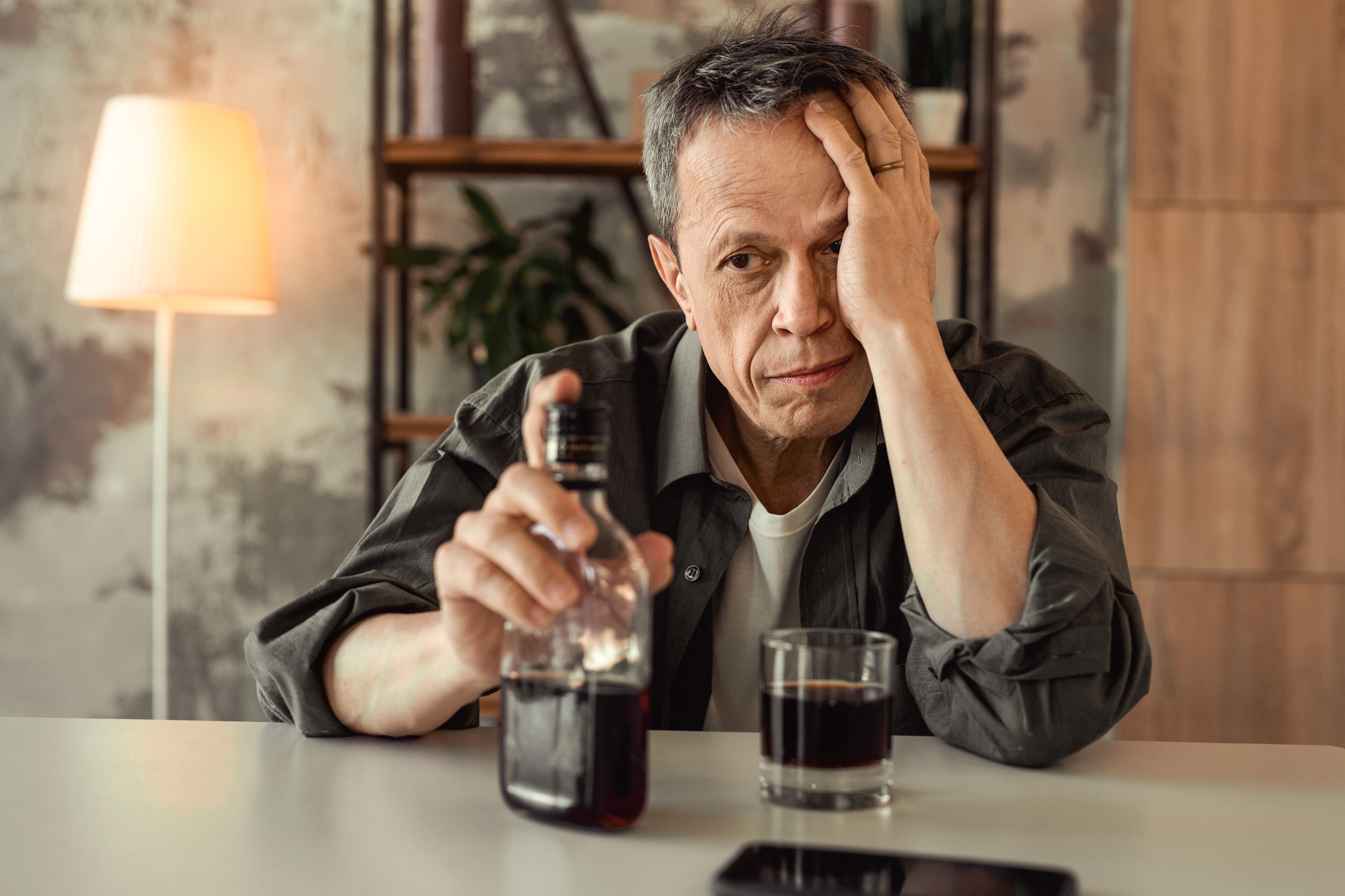 uomo maturo con bottiglia e bicchiera di alcolico, una mano sulla faccia e l'espressione stanca, concept: Alcol Anziani