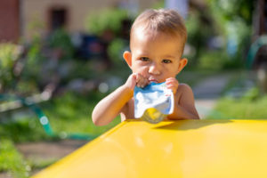 bambino serio che mangia purea di frutta nel sacchetto e guarda nella telecamera davanti al tavolo giallo. sullo sfondo c'è un giardino verde in una giornata di sole sfocato