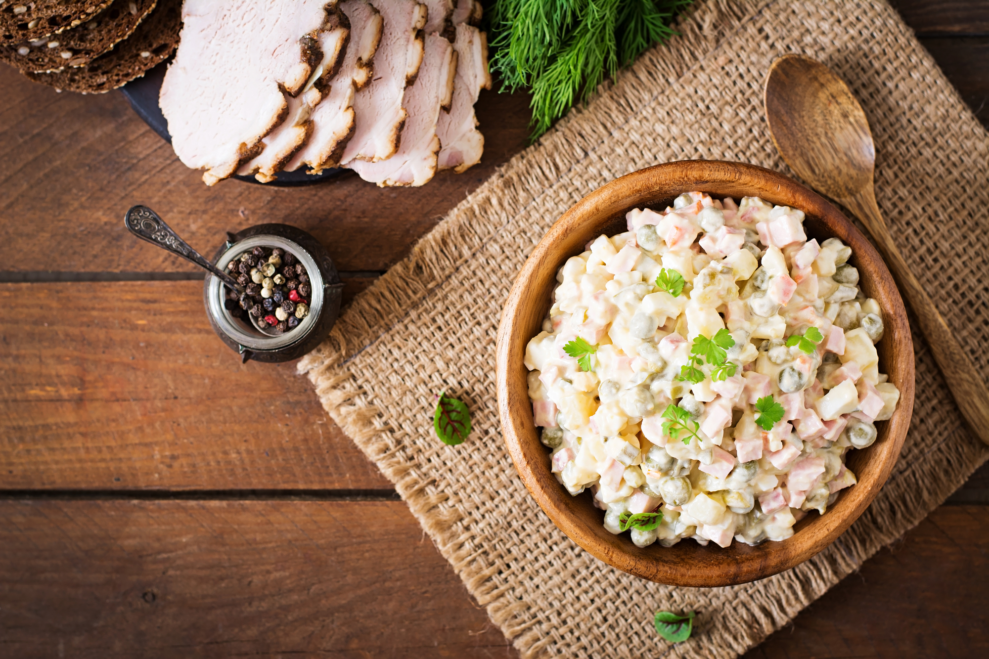 Insalata russa presentata in piatto di legno su tela di iuta su tavolo di legno con vasetto di pepe e carne affettate e fette di pani