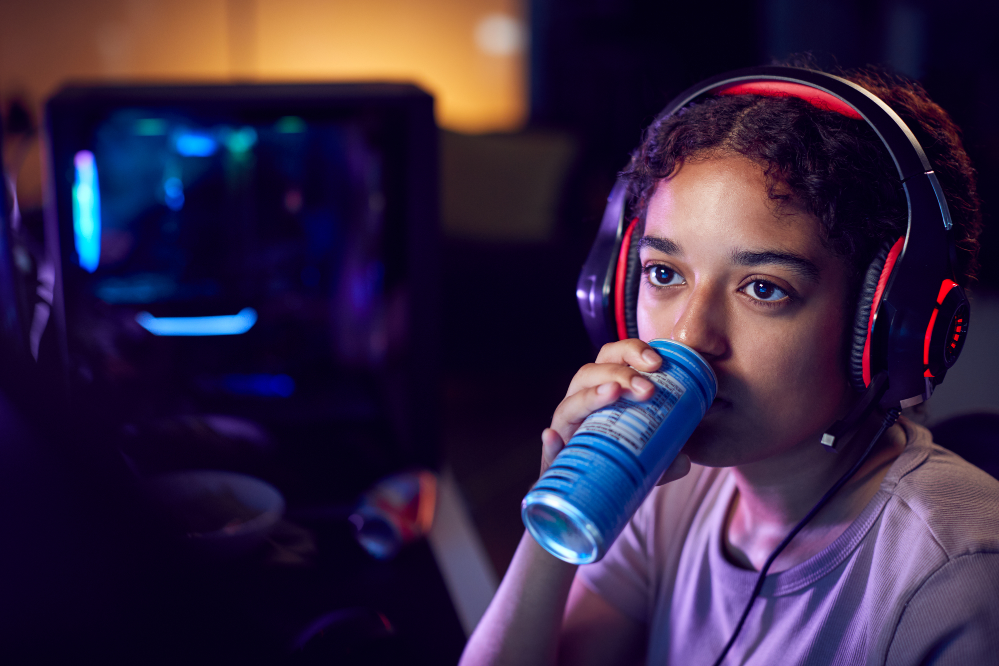 Ragazza adolescente con cuffie da gaming beve bevanda zuccherata o energy drink alla scrivania davanti al computer