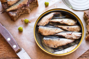 Sardine sott'olio in scatola, accanto a sardine su fetta di pane integrale, coltello, alcuni piselli sparsi