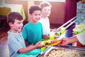 Bambini in fila al bancone della mensa scolastica mentre un'operatrice passa un piatto con carote, broccoli e pasta al primo della fila