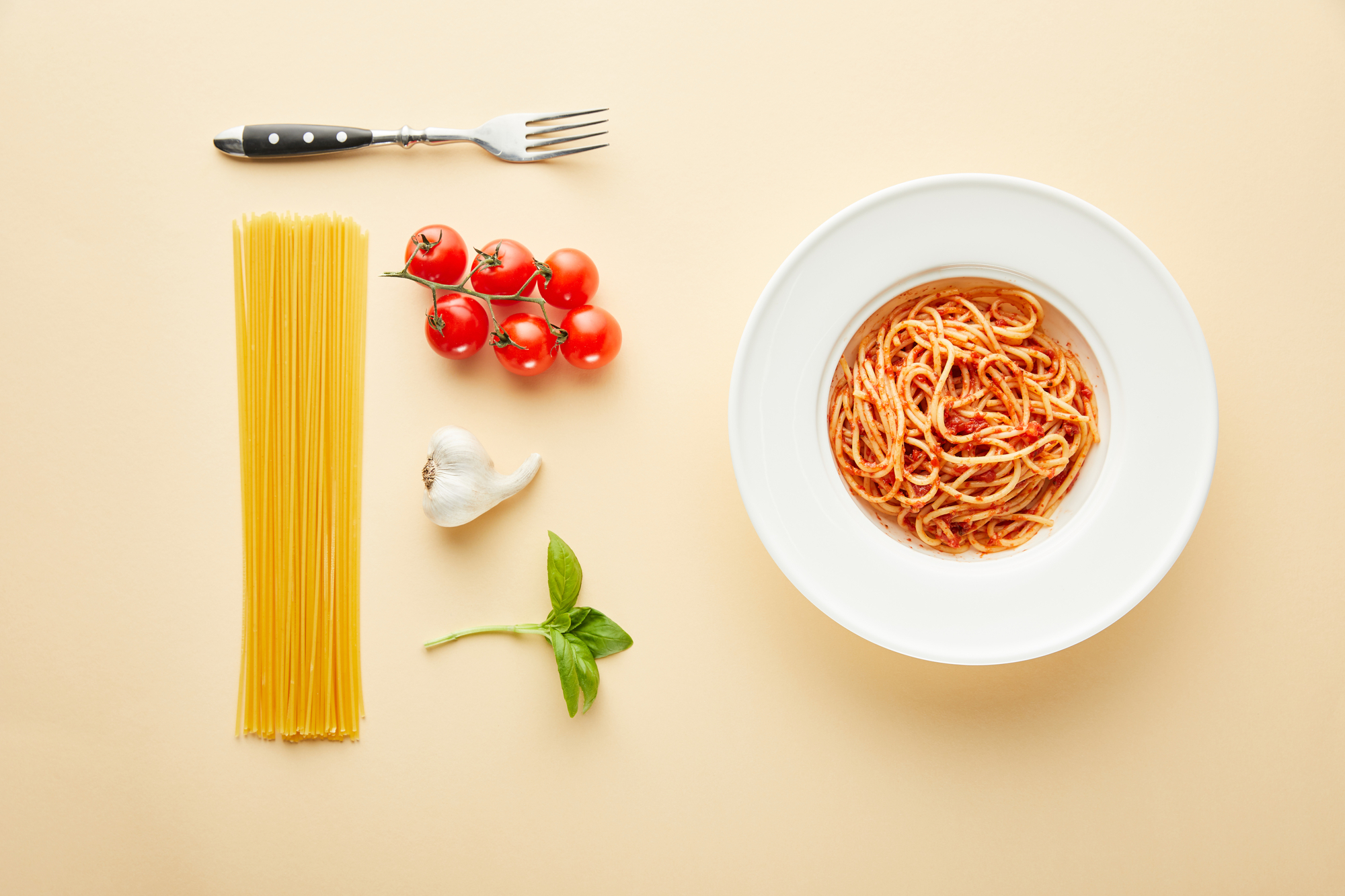 Piatto di spaghetti al pomodoro; sulla sinistra gli ingredienti: spaghetti crudi, pomodorini, aglio, basilico e una forchetta
