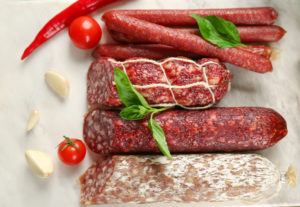Tre salami tagliati e alcuni salamini interi su uno sfondo bianco con foglie di basilico, pomodorini e aglio