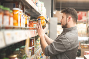 Uomo esamina etichetta di una conserva vegetale in vaso di vetro davanti agli scaffali del supermercato