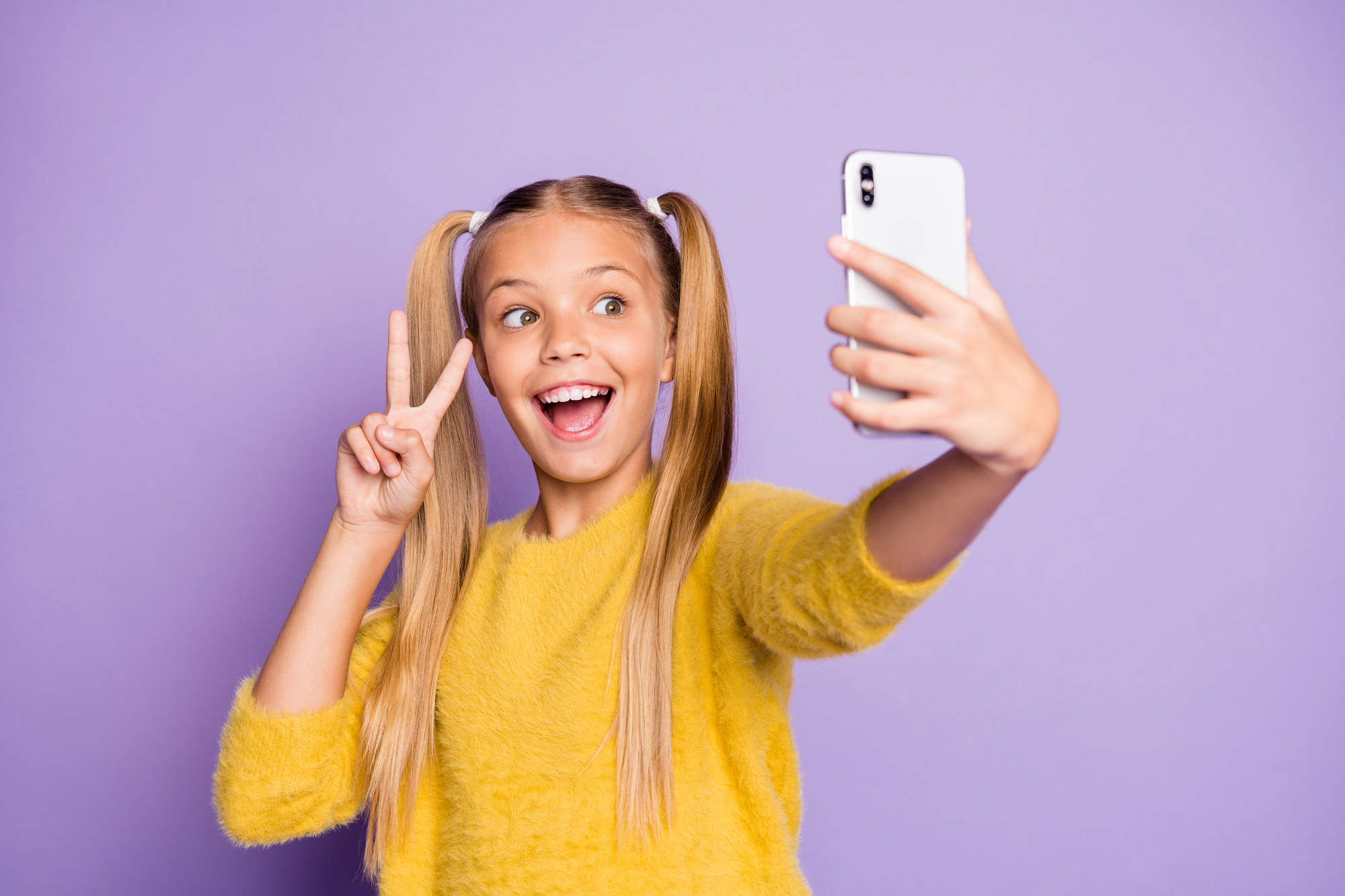 Bambina sorridente che fa il segno di vittoria mentre si fa un selfie con lo smartphone; concept: baby influencer, social media
