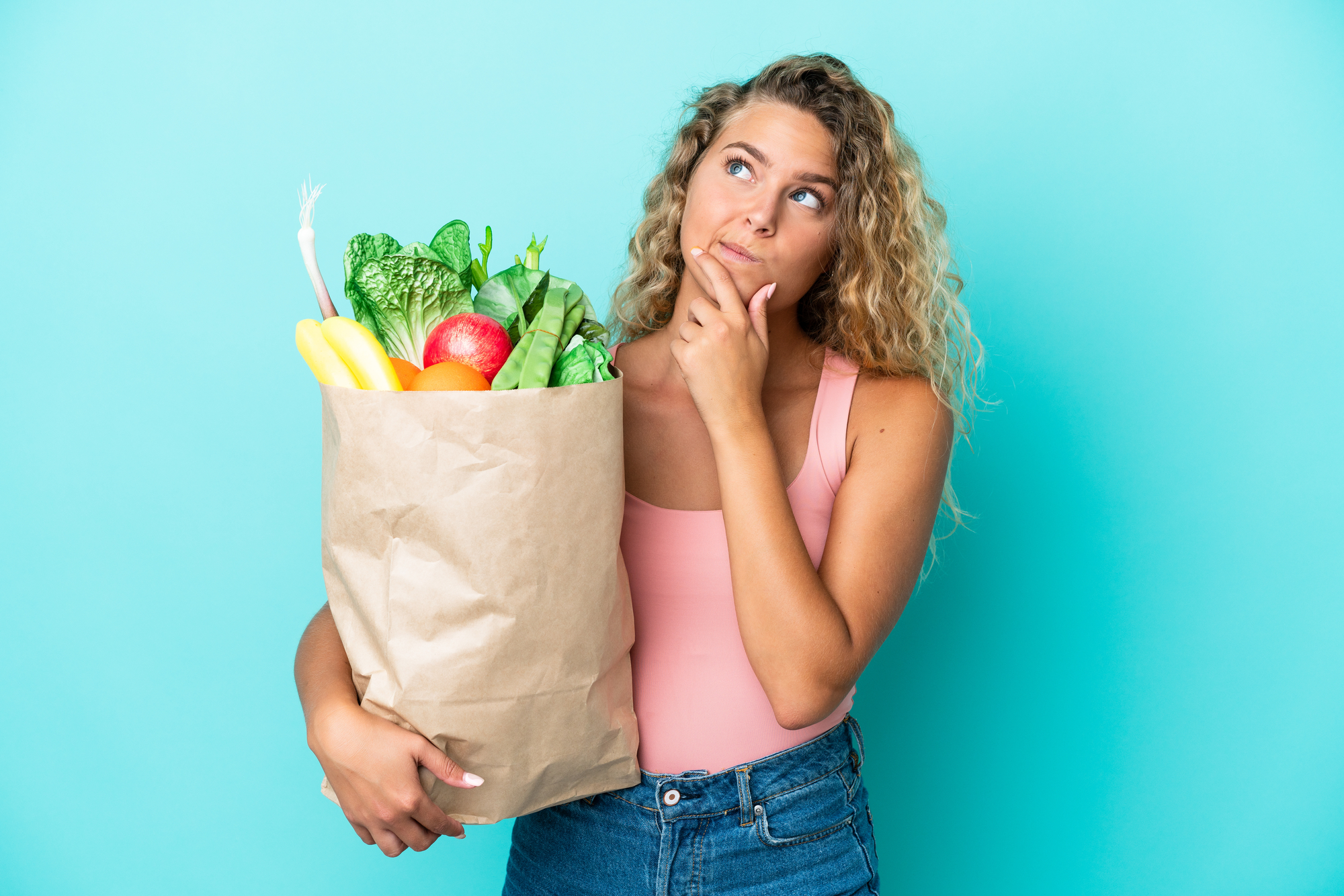 Ragazza con i capelli ricci che tiene una busta della spesa isolata su sfondo verde che ha dei dubbi supermercato donna frutta e verdura diete perplessità