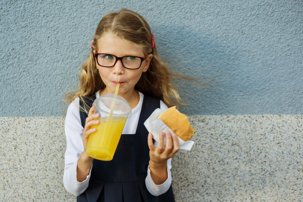 Bambina con hamburger in mano beve aranciata con la cannuccia da bicchiere di plastica grande; junk food