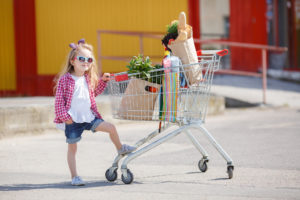 Bambina con occhiali da sole appoggiata a carrello con la spesa nel parcheggio di un supermercato