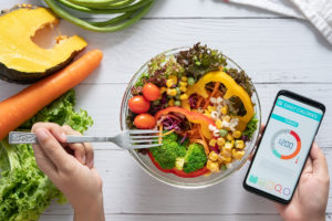 Una persona mangia un'insalata di cereali, verdure e legumi, mentre tiene in mano uno smartphone con app conta calorie; concept: dieta